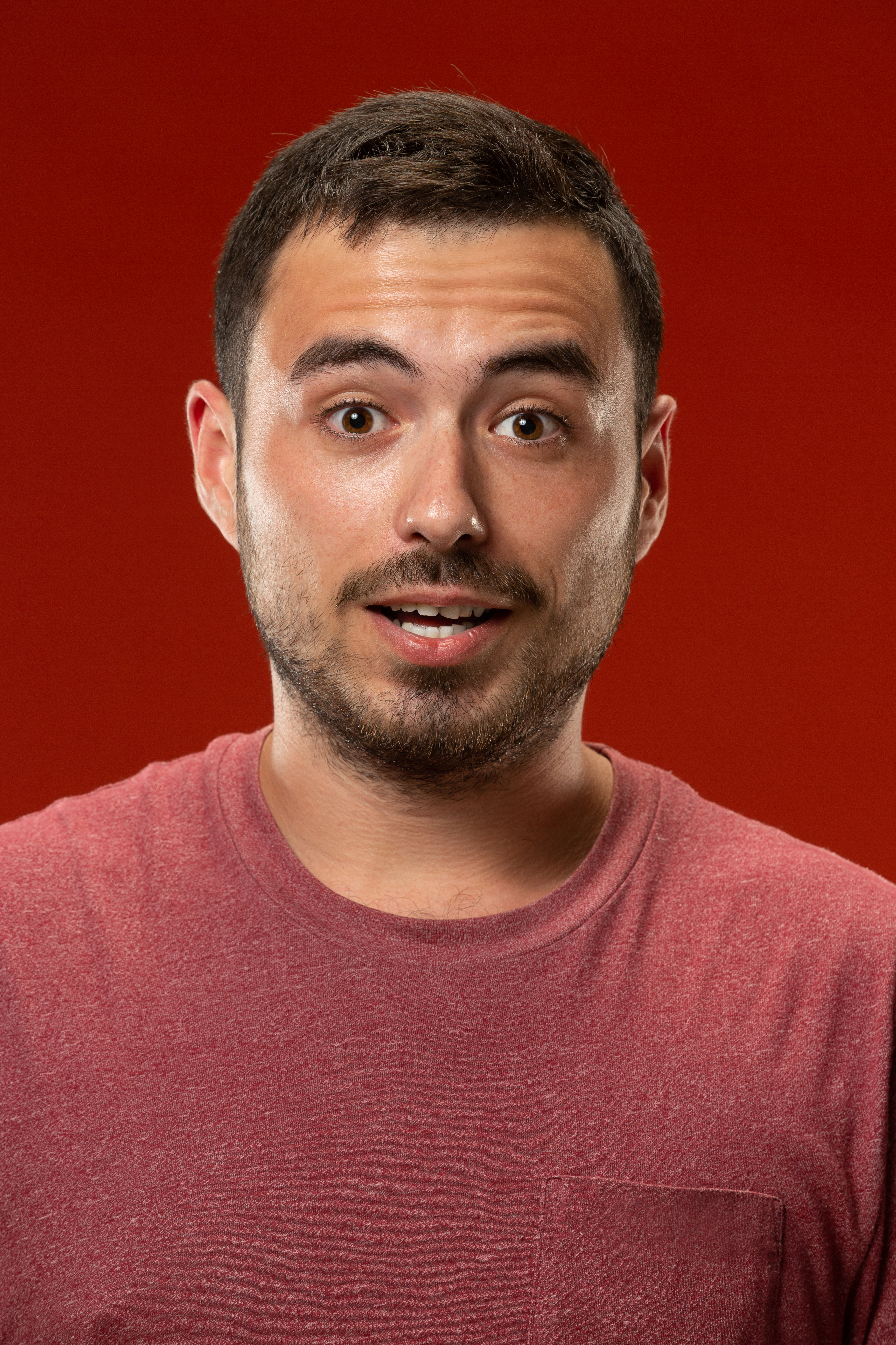 Wow. Attractive male half-length front portrait on red studio backgroud. Young emotional surprised bearded man standing with open mouth. Human emotions, facial expression concept. Trendy colors